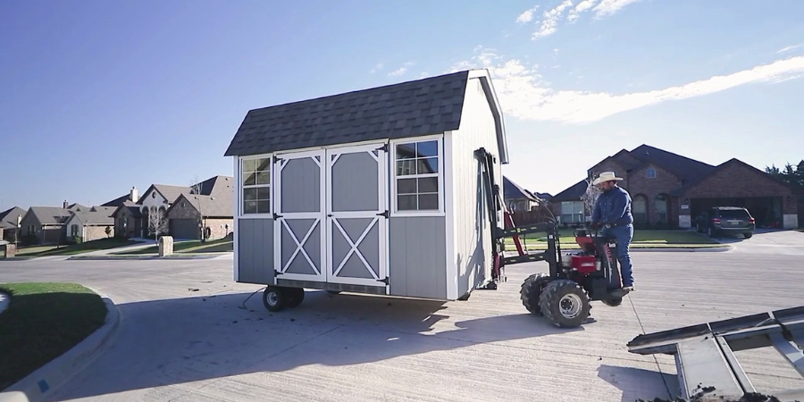 Worker in a cowboy hat maneuvering a gray gambrel-roof storage building into place with a shed mule on a suburban street