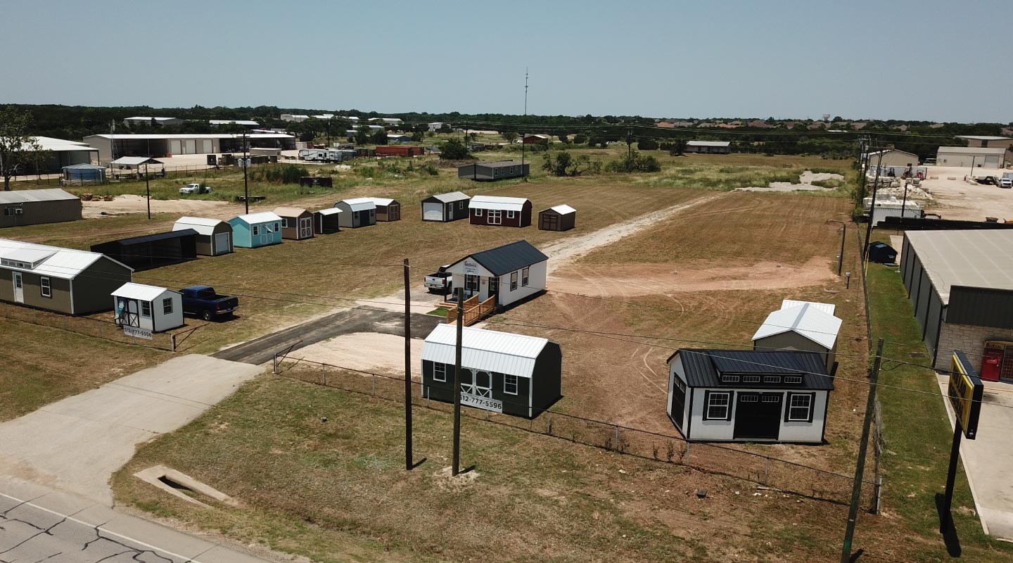 Aerial view of the Quality Storage Buildings display lot in Liberty Hill, with two rows of storage sheds and lofted barns, a white sales office, and gravel drives off the highway