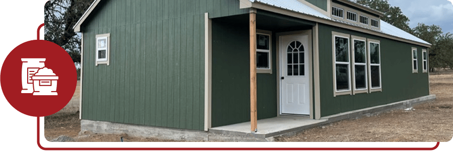 Green cabin with a covered porch built on a poured concrete pad, with the slab edge visible along the base of the walls