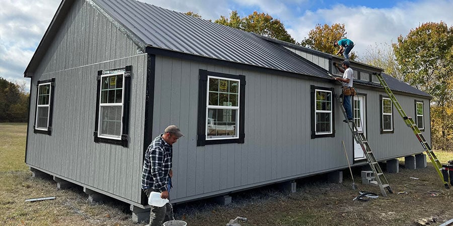 Two workers setting a gray cabin-style building onto concrete block piers in an open field, one on a ladder finishing the roofline