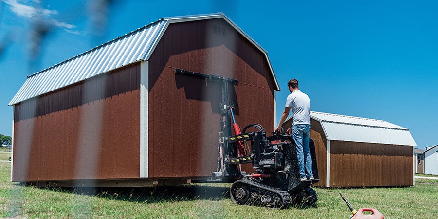 Worker on a tracked shed mule repositioning a dark red gambrel-roof barn on a grass lot, with a second brown barn behind it