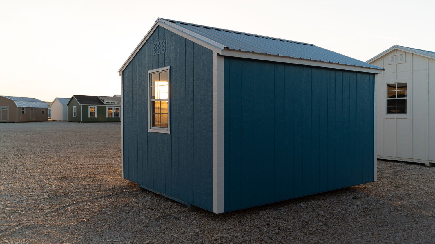 Navy blue garden shed with white trim and a metal gable roof on a gravel lot at sunset, with more storage buildings lined up behind it