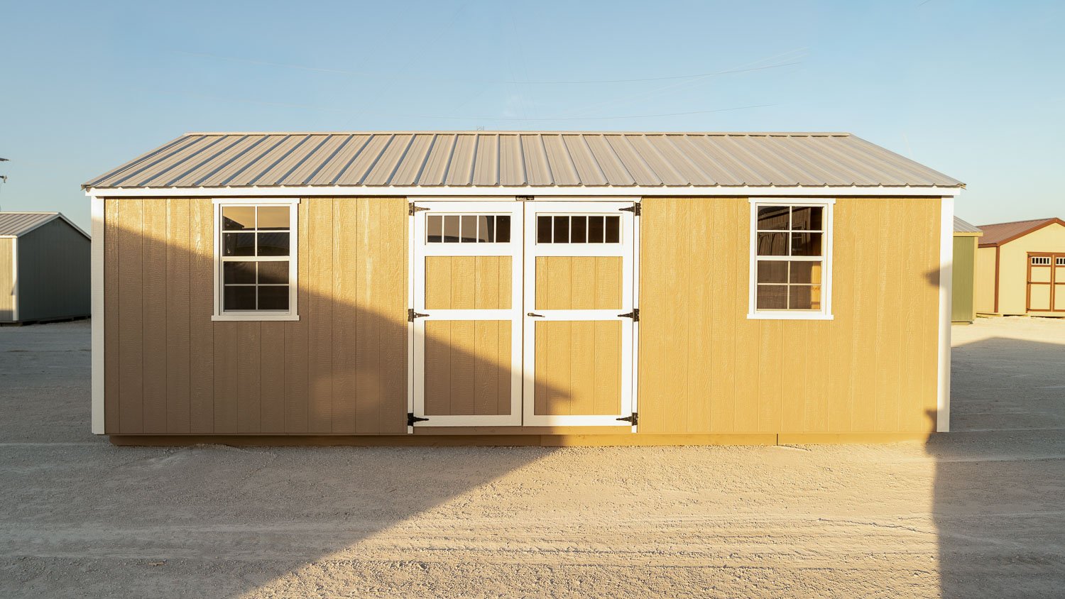 Buttercream-yellow garden shed with white trim, white double doors with transom glass, and a tan standing-seam metal roof on a gravel lot