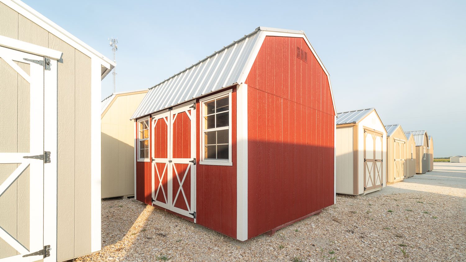 Red lofted barn with a gambrel roof and white double barn doors on a display lot beside other storage buildings