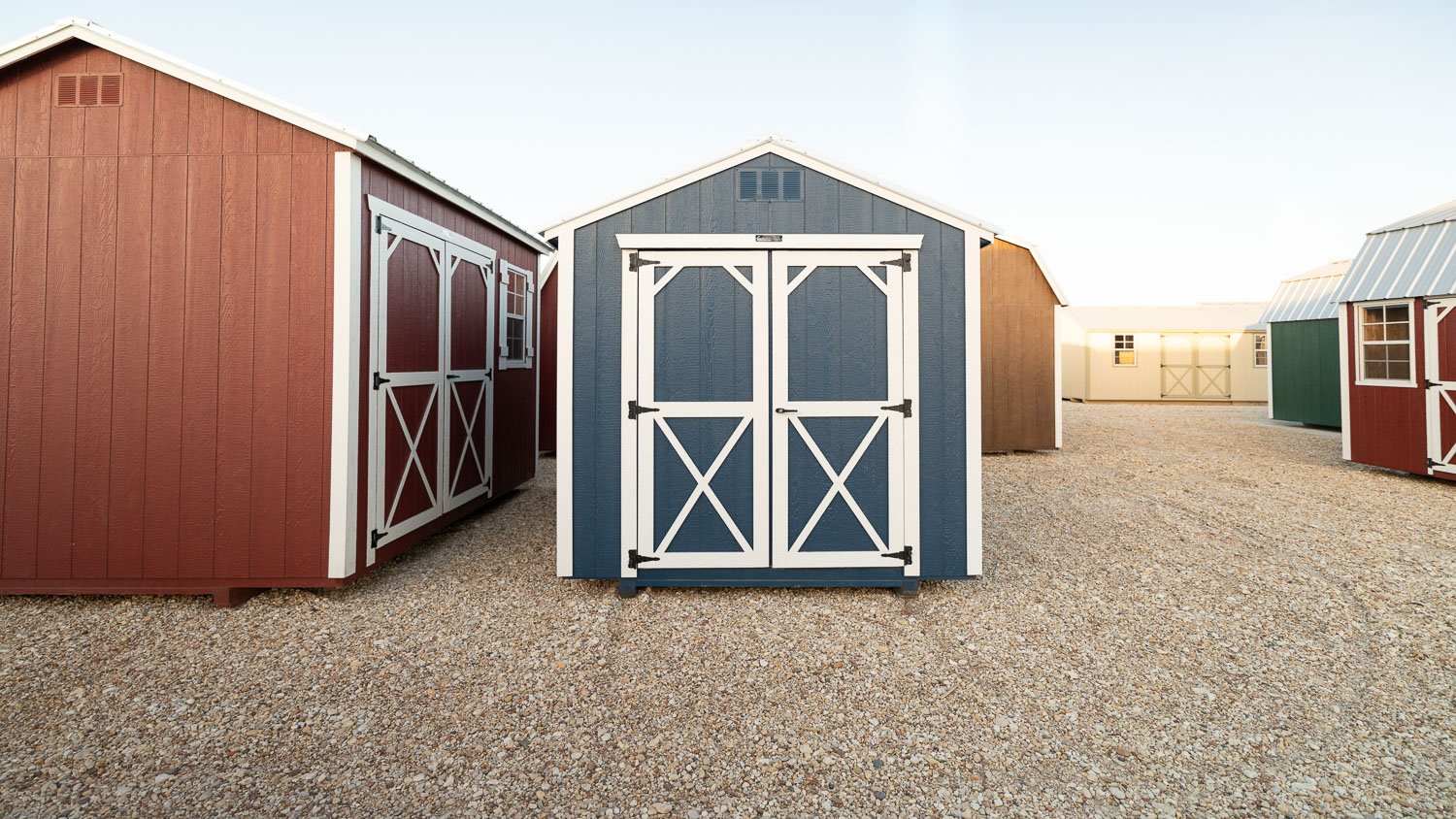Blue-gray utility shed with white cross-braced double doors on a gravel display lot, with red and tan storage buildings on either side