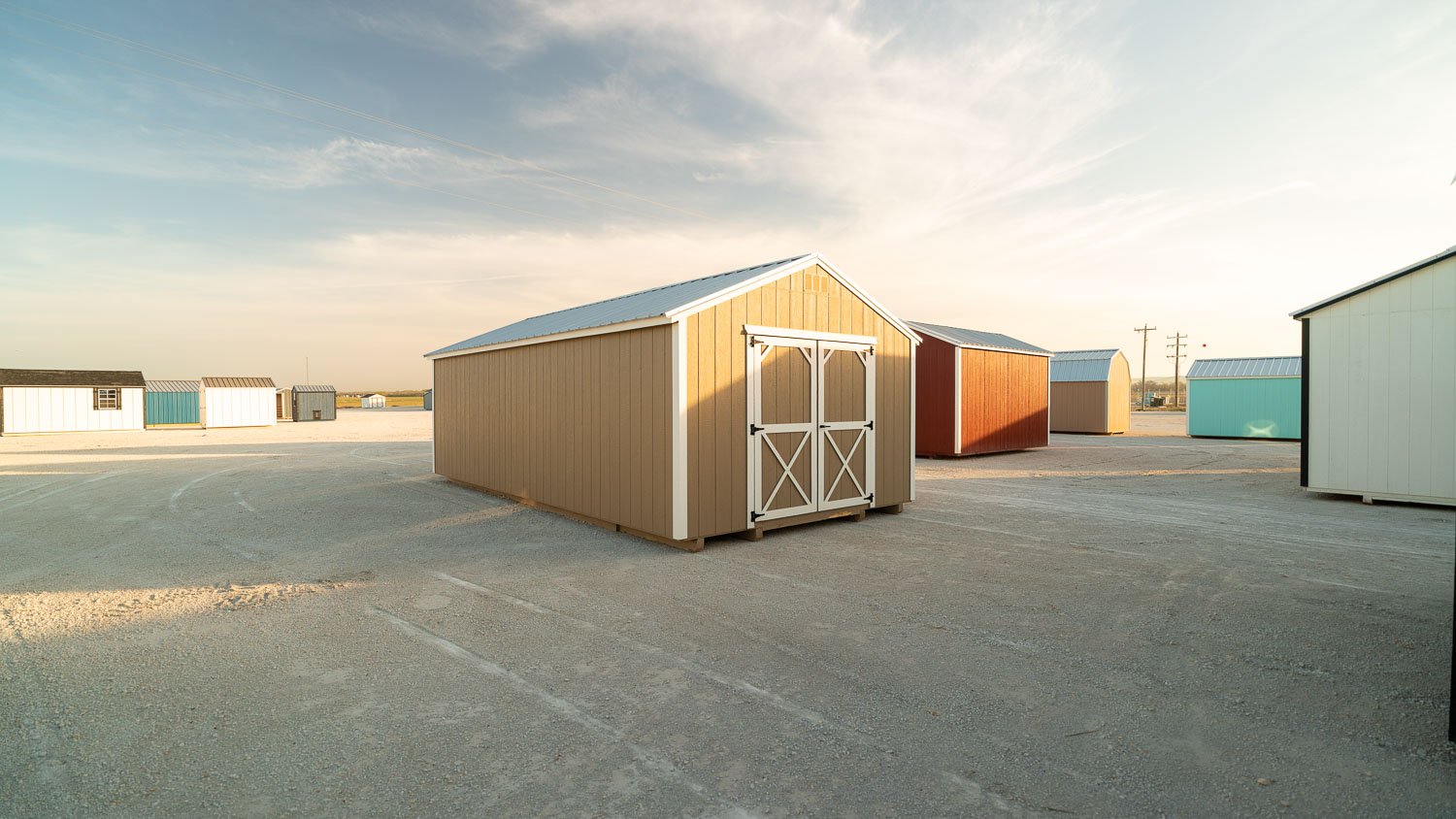 Khaki utility shed with white trim and white X-braced double doors under a blue metal roof, with white, red, teal, and brown buildings in other colors around it