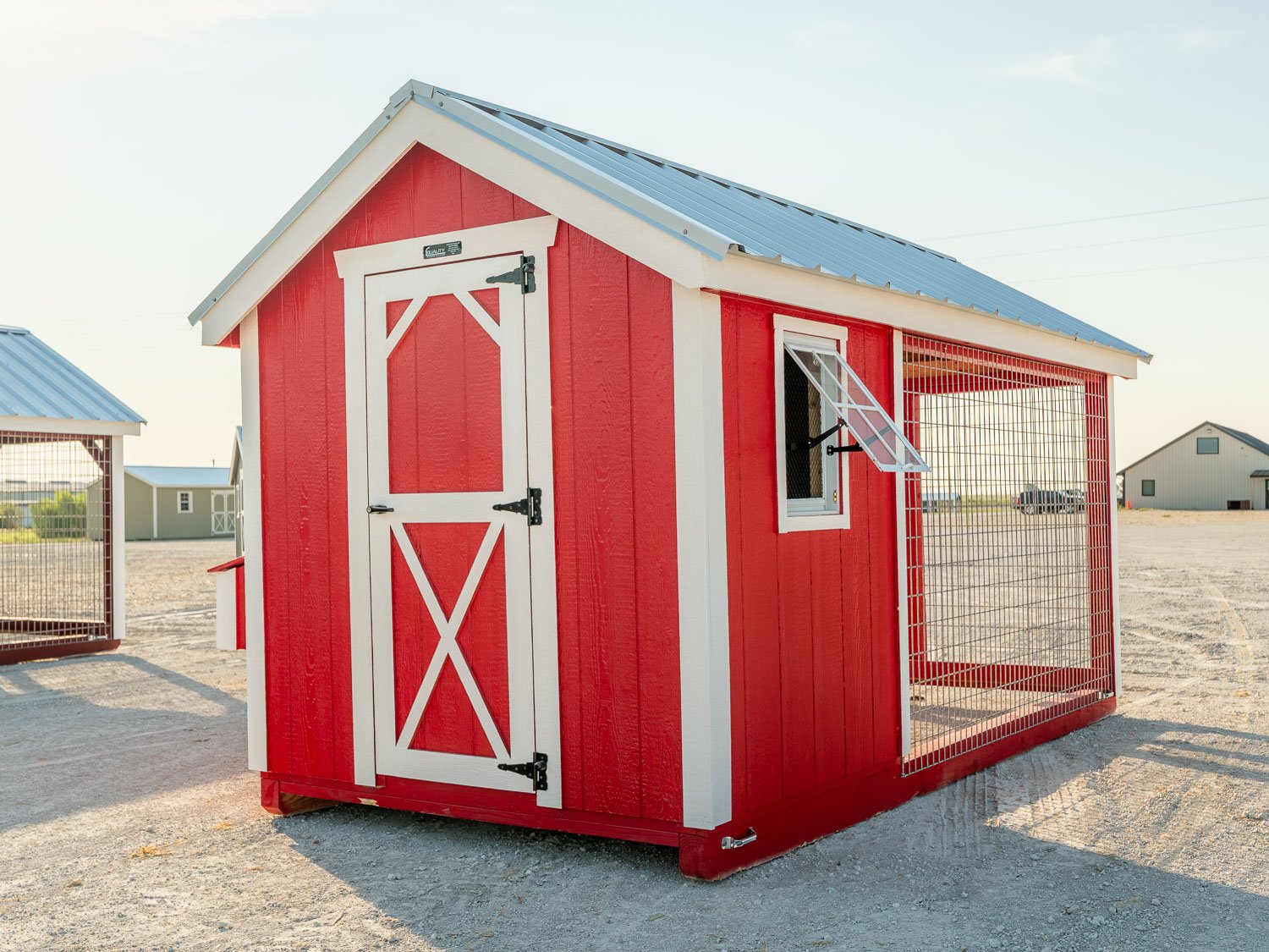 Red chicken coop with attached wire-mesh run and an open awning window, sitting on a gravel lot with other storage buildings in the background