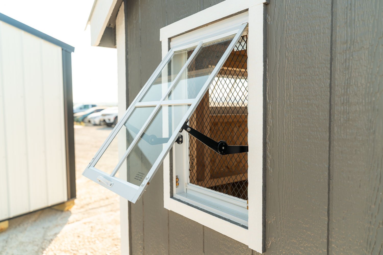 Close-up of a coop's open awning window showing the wire mesh screen, hinge arm, and hardware that provide ventilation