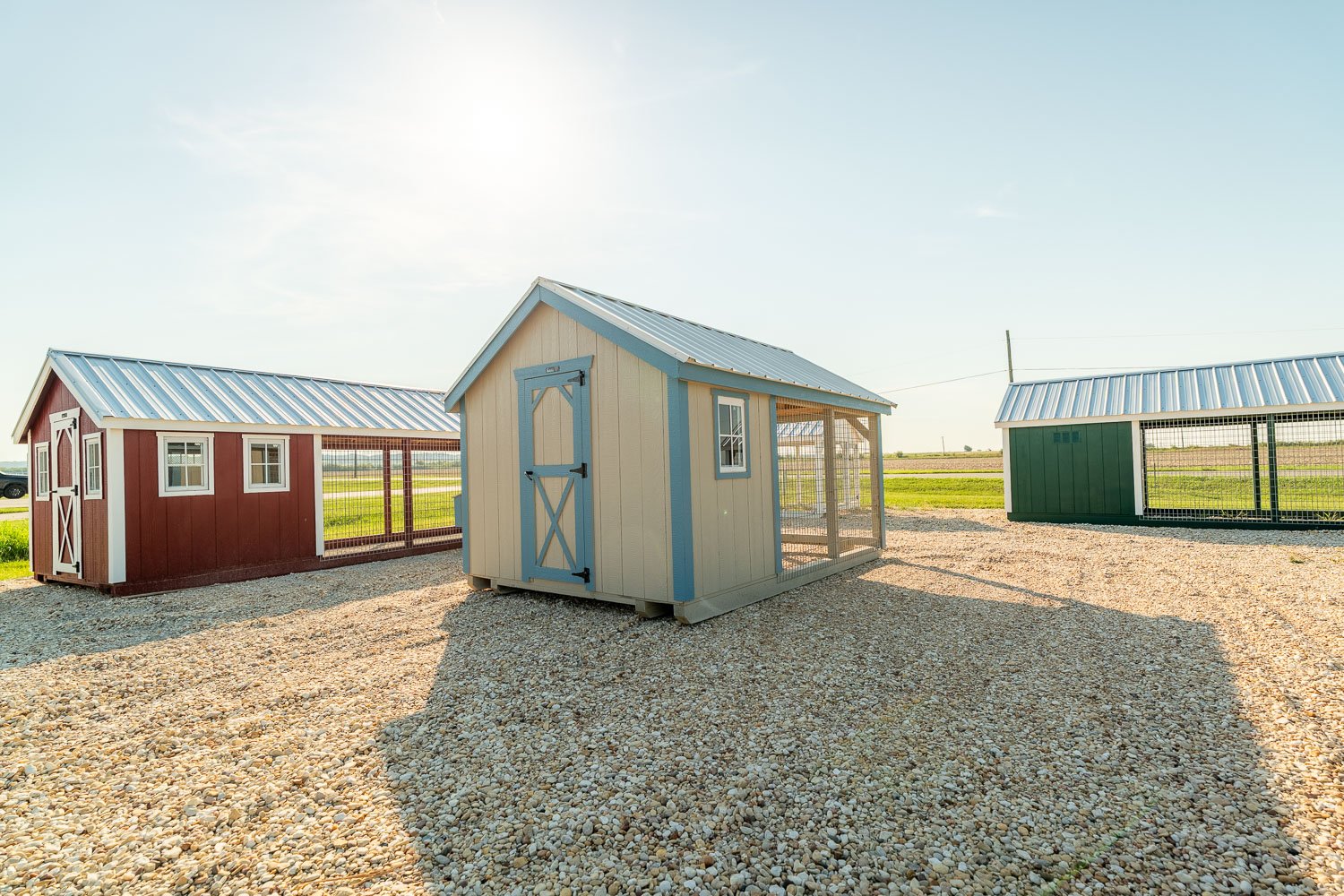 Three chicken coops in red, tan, and green, each with an attached wire-mesh run, displayed side by side on a gravel lot