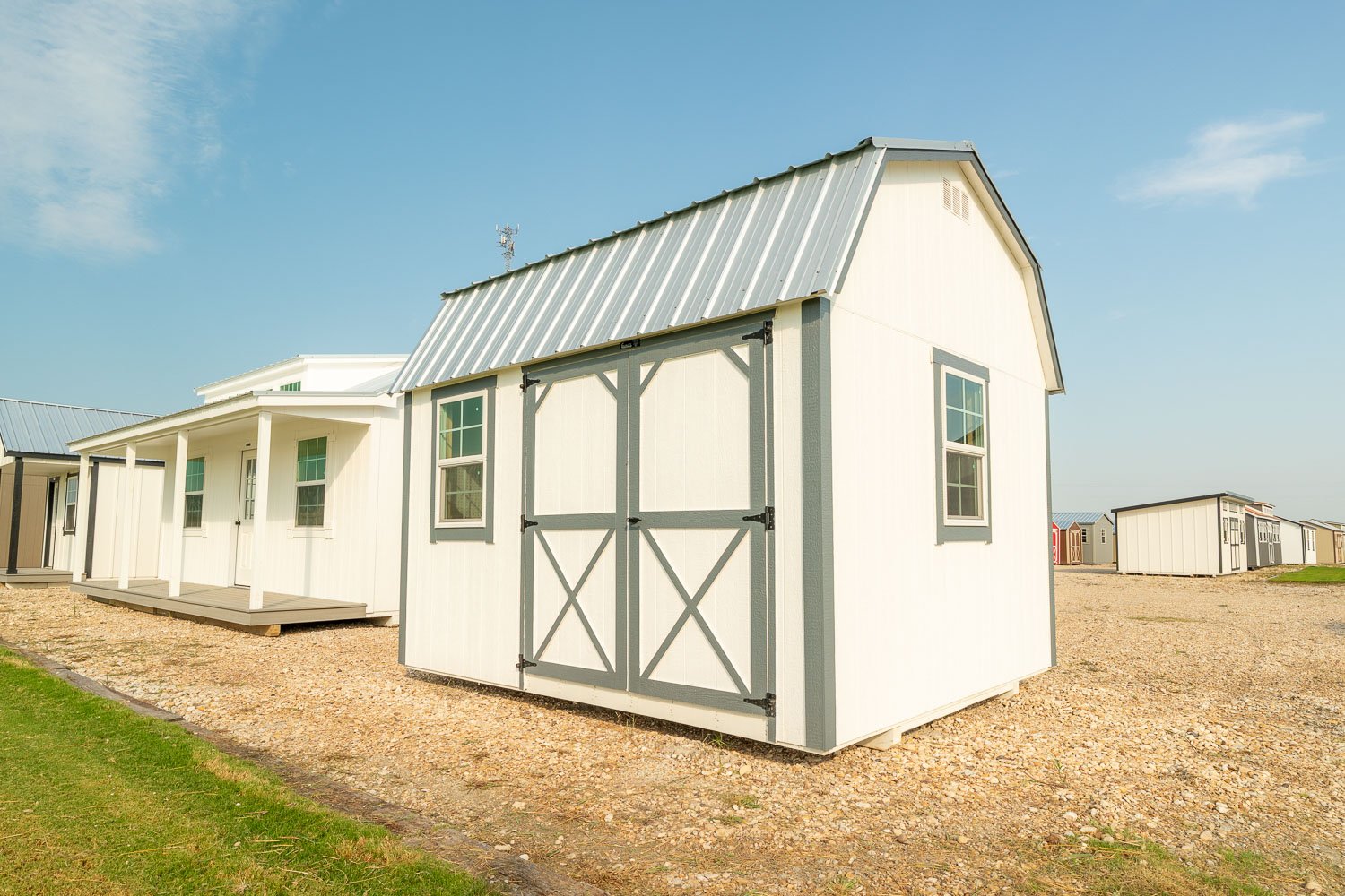 Cream lofted barn with a gray gambrel metal roof and gray cross-braced double doors on a gravel lot, with a white porch cabin behind it