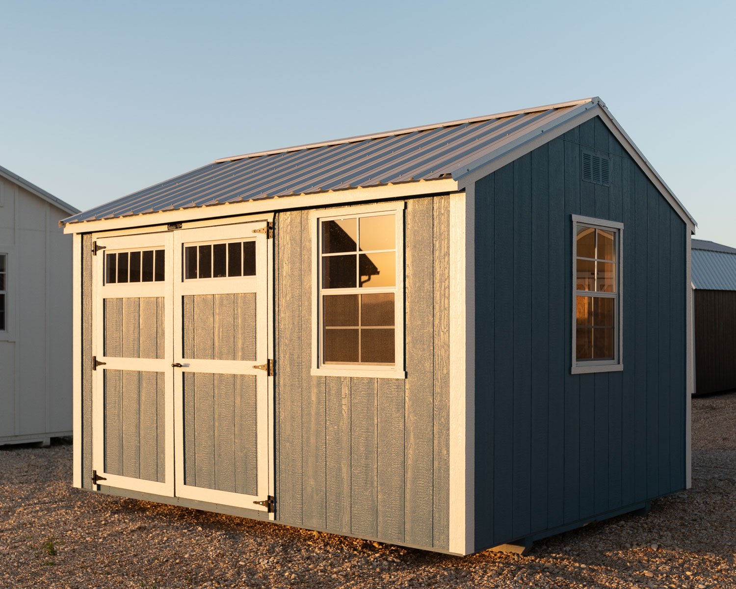 Blue-gray gable-roof garden shed with white trim, double doors with transom windows, a window beside the door, and a gable-end window, viewed at an angle on a gravel lot under warm, low-angle sunlight