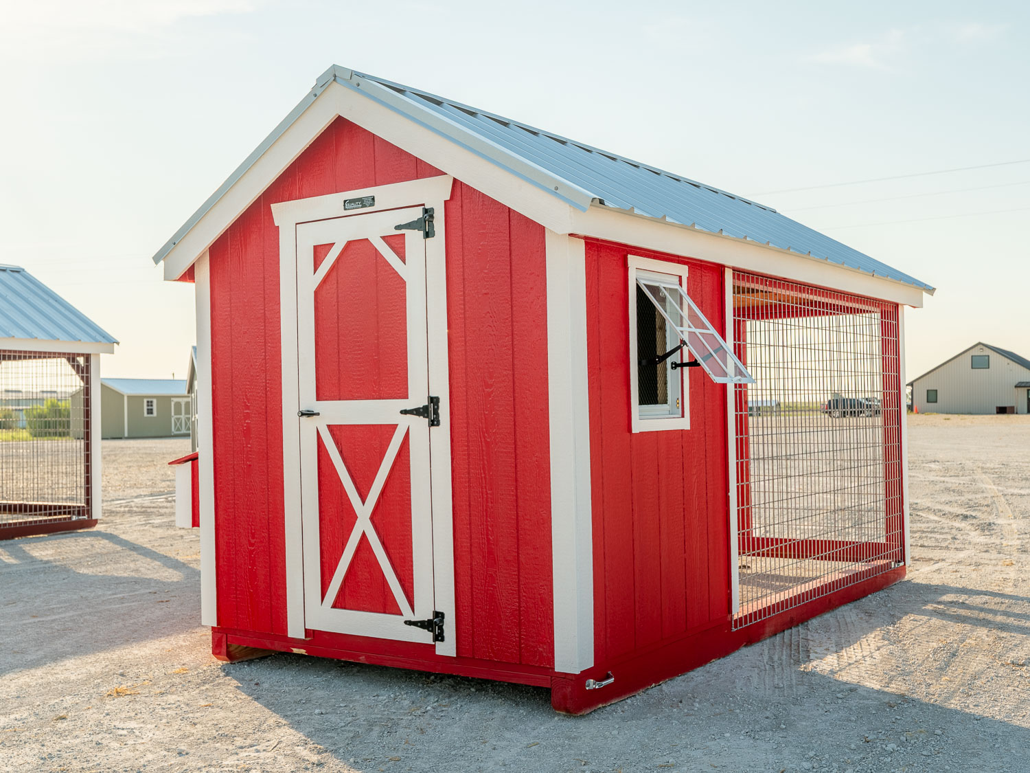 Red chicken coop with attached wire-mesh run and an open awning window, sitting on a gravel lot with other storage buildings in the background