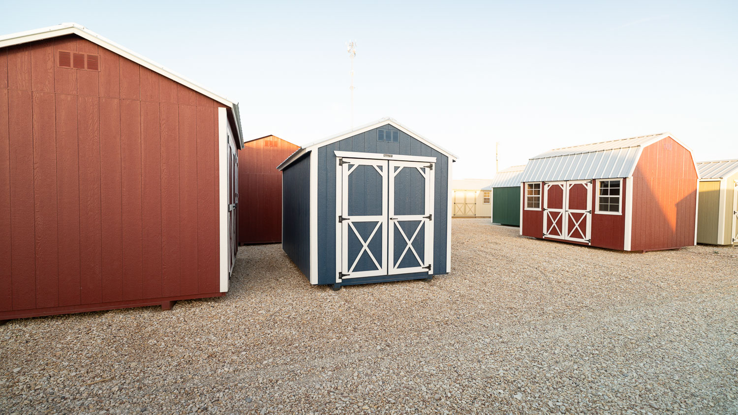 Blue gable-roof utility shed with white trim, solid X-braced double doors with no windows, and a louvered vent on the gable wall, sitting among red barn-style and gable sheds at varying angles on a gravel display lot
