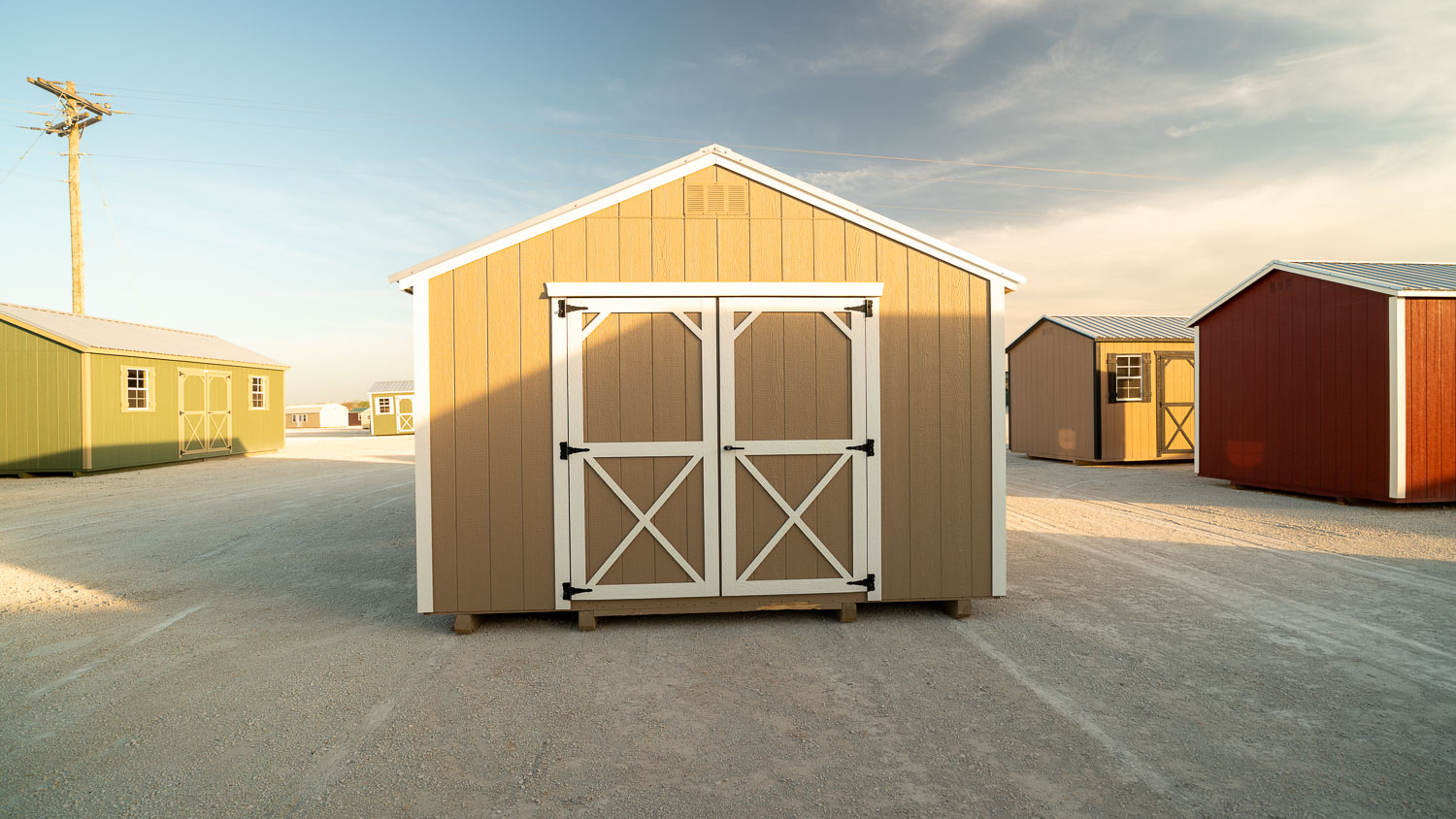 Tan utility shed with white trim, gable vent, and white cross-braced double doors, sitting on wood skids on a gravel lot